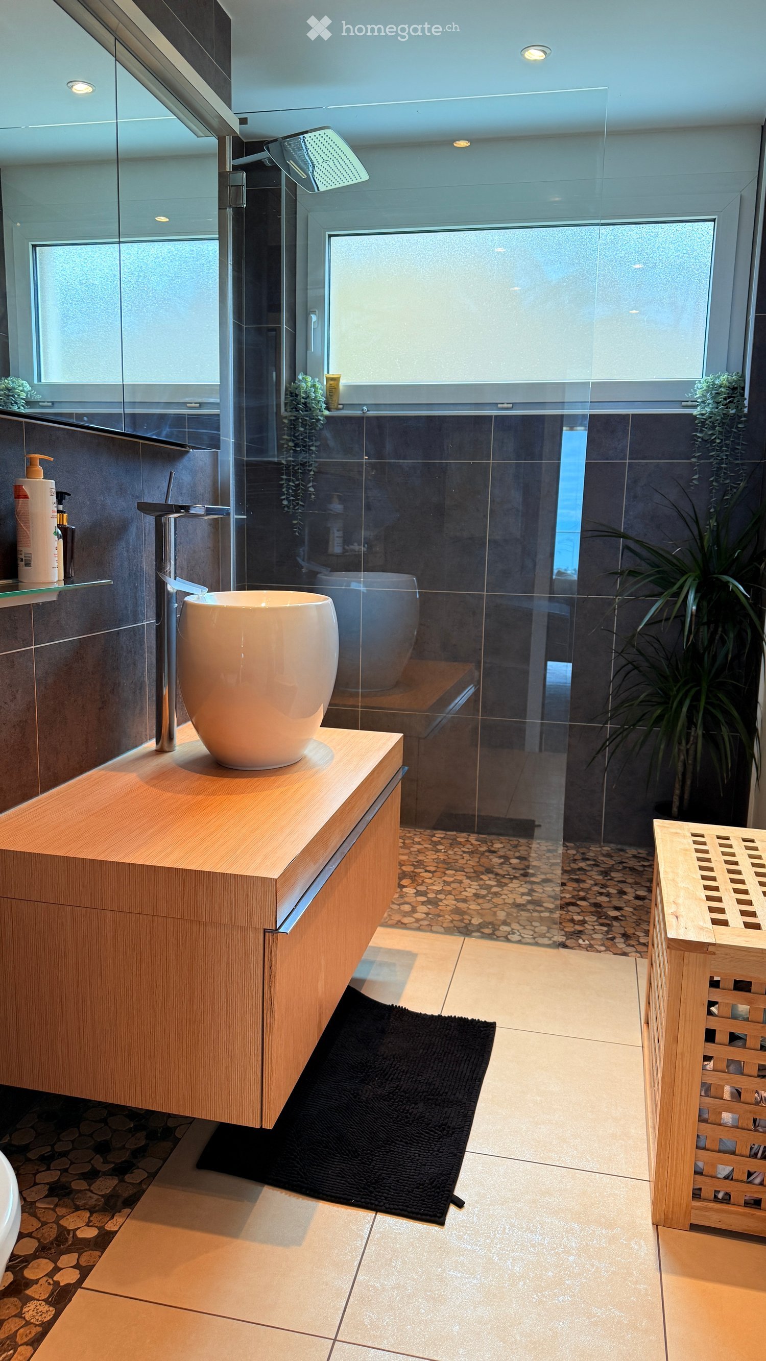 Modern bathroom with a wooden vanity, large sink, glass-enclosed shower, and dark tiled walls.