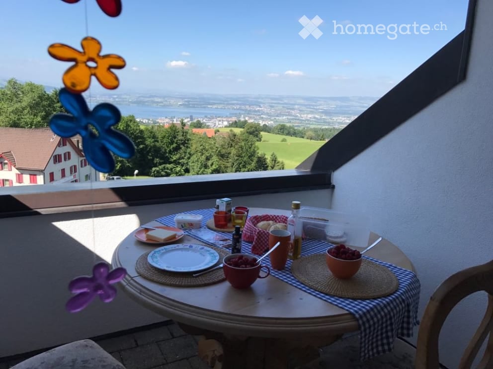 Balcony with a round table set for breakfast, overlooking a scenic landscape with a lake and distant town.