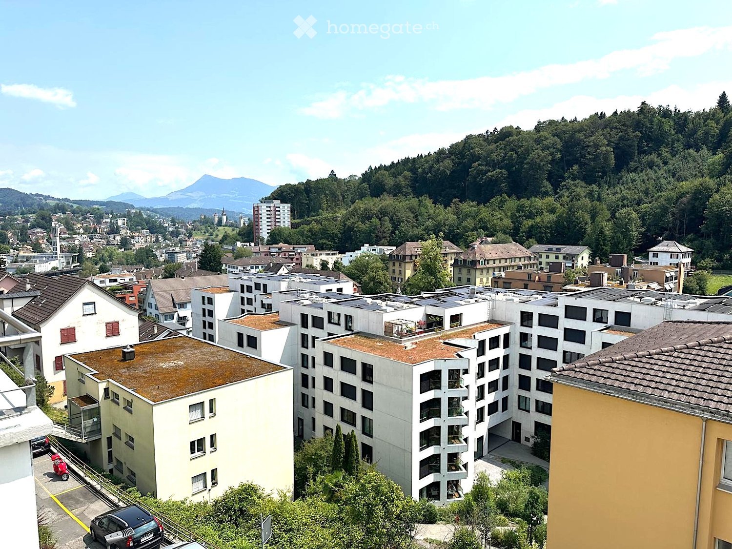 Aerial view of a residential area with multiple apartment buildings, surrounded by greenery and distant mountains.