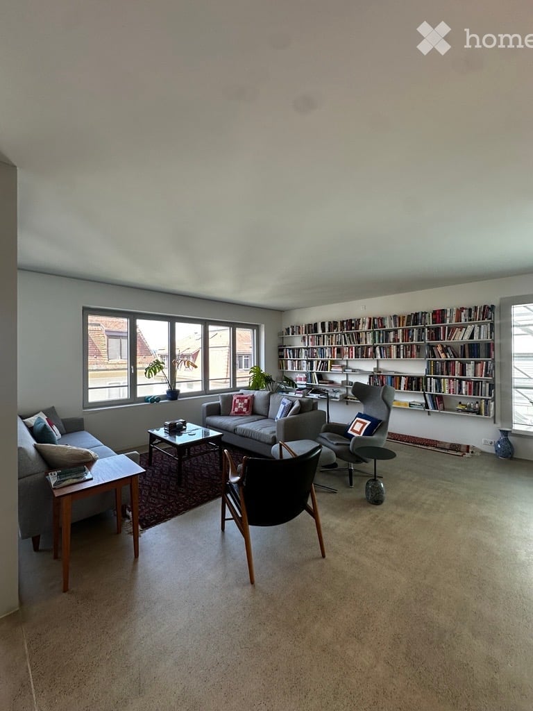 Bright living room with large windows, grey sofa, coffee table, bookshelf, and decorative plants.
