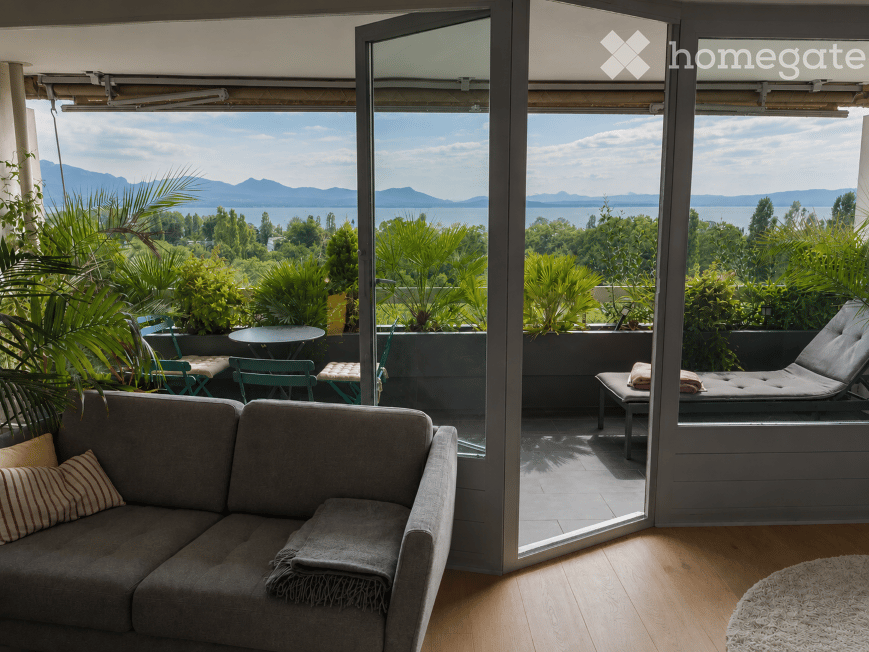 Living room with a grey sofa, wooden floor, large windows opening to a balcony with outdoor furniture and scenic mountain view.