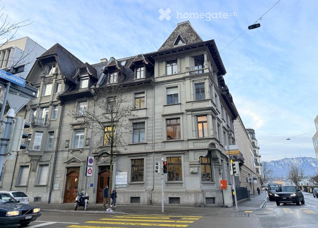 A multi-story building with a classic facade, large windows, and ornate architectural details. The building has a dark roof with dormer windows and is situated on a street corner with traffic signals and a pedestrian crossing.