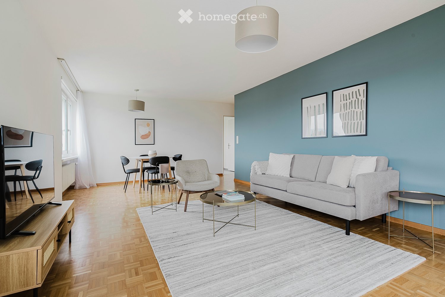 Modern living room with a grey sofa, coffee table, armchair, dining area, TV stand, and artwork on the walls.