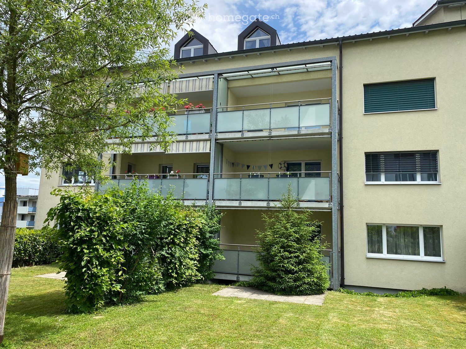 Three-story building with multiple balconies, beige exterior walls, green grass and plants, clear sky