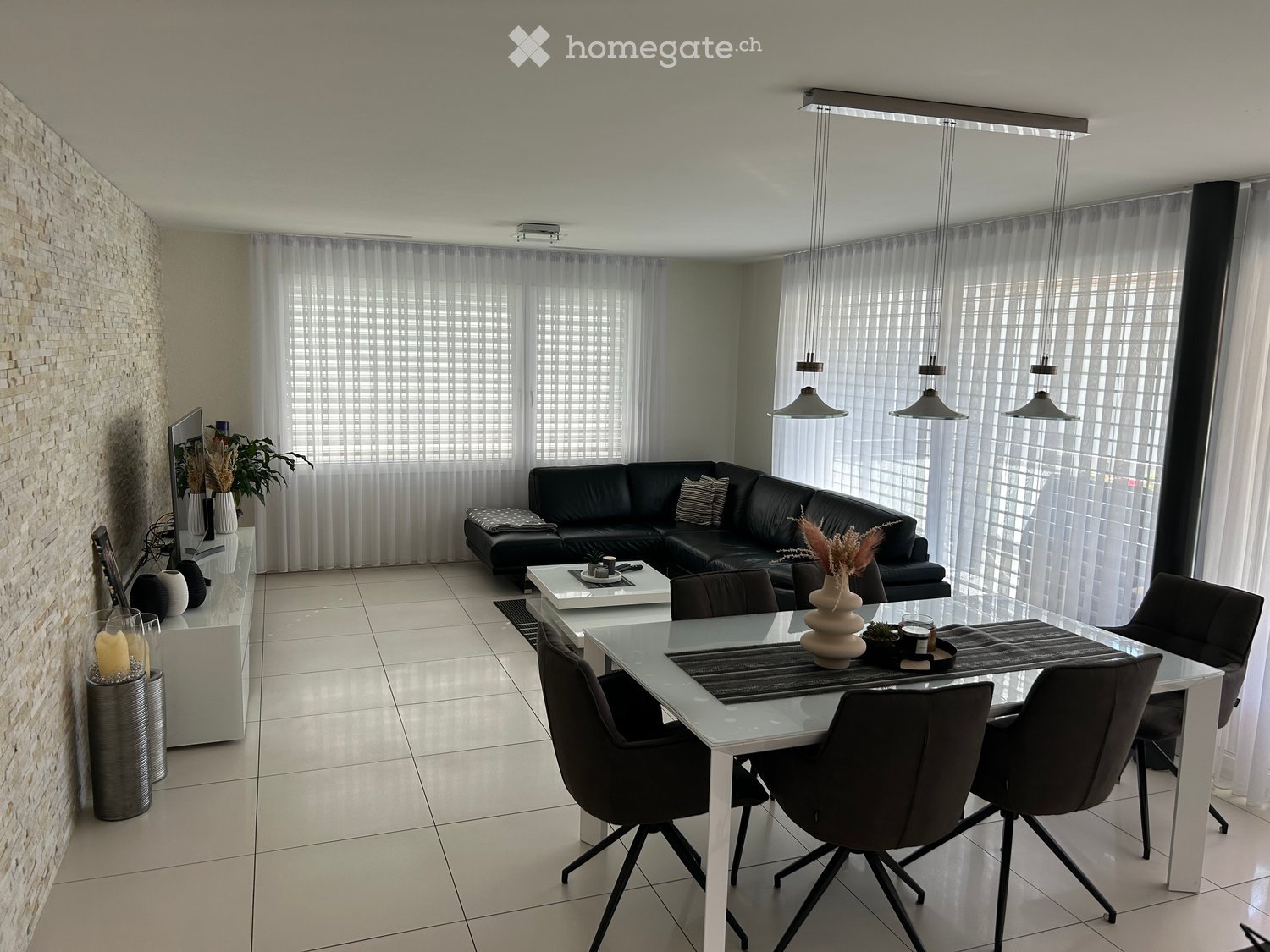 Modern living and dining area with a black leather sofa, white coffee table, glass dining table with chairs, decorative vases, and a TV screen.