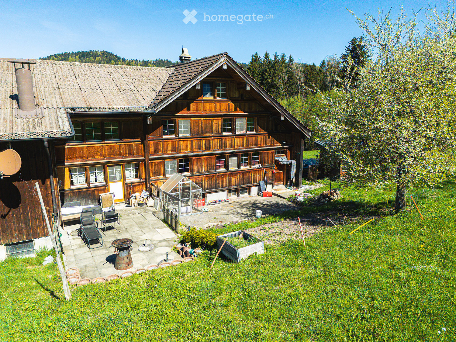 Traditional wooden house with a sloped roof, multiple windows, and a spacious garden area with outdoor furniture and a barbecue pit.