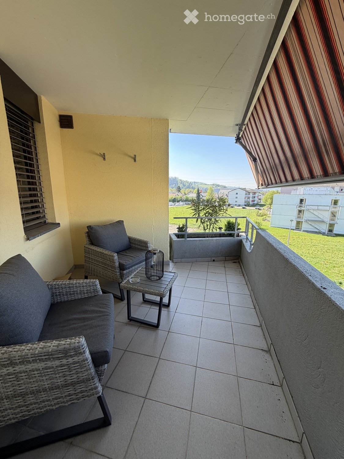 Balcony with tiled floor, wicker sofa, wicker chair, small table, and a view of the surrounding area.