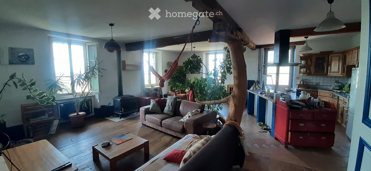 A cozy living room with a wooden floor, large windows, a fireplace, a sofa, coffee table, and a kitchen area with cabinets and appliances.