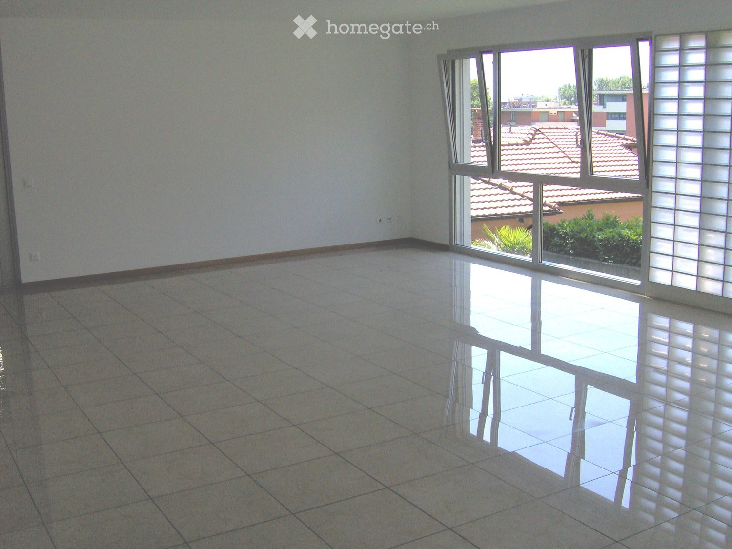 Empty room with tiled floor and large windows showing a view of neighboring buildings and greenery.