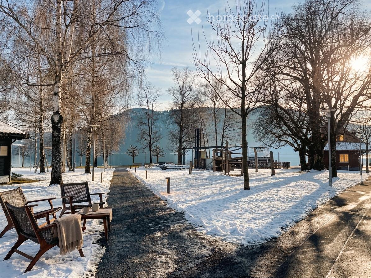 A snow-covered pathway lined with trees leads to a wooden bench and table setup with a cup and blanket, surrounded by bare trees and a distant mountain view.