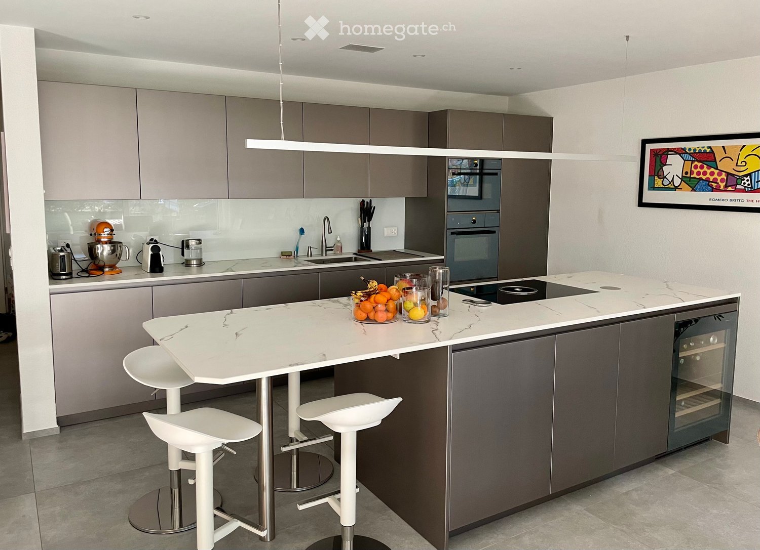 Modern kitchen with grey cabinets, white island, marble countertop, four white stools, built-in oven, sink, and various appliances.
