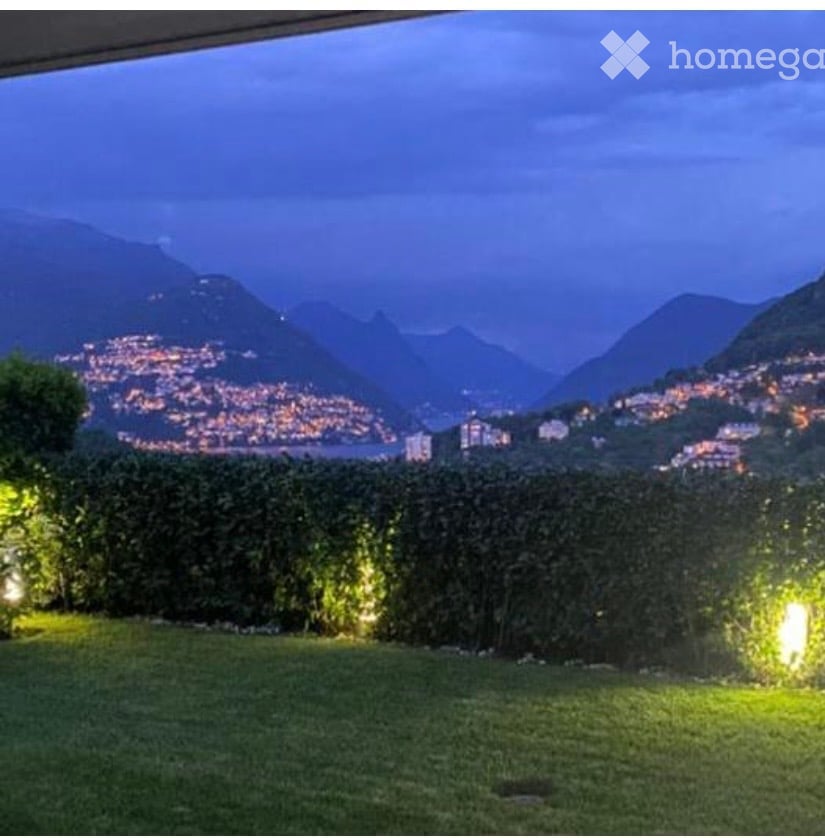 Evening view of a garden with well-lit hedges and a cityscape in the background, featuring mountains and illuminated buildings.