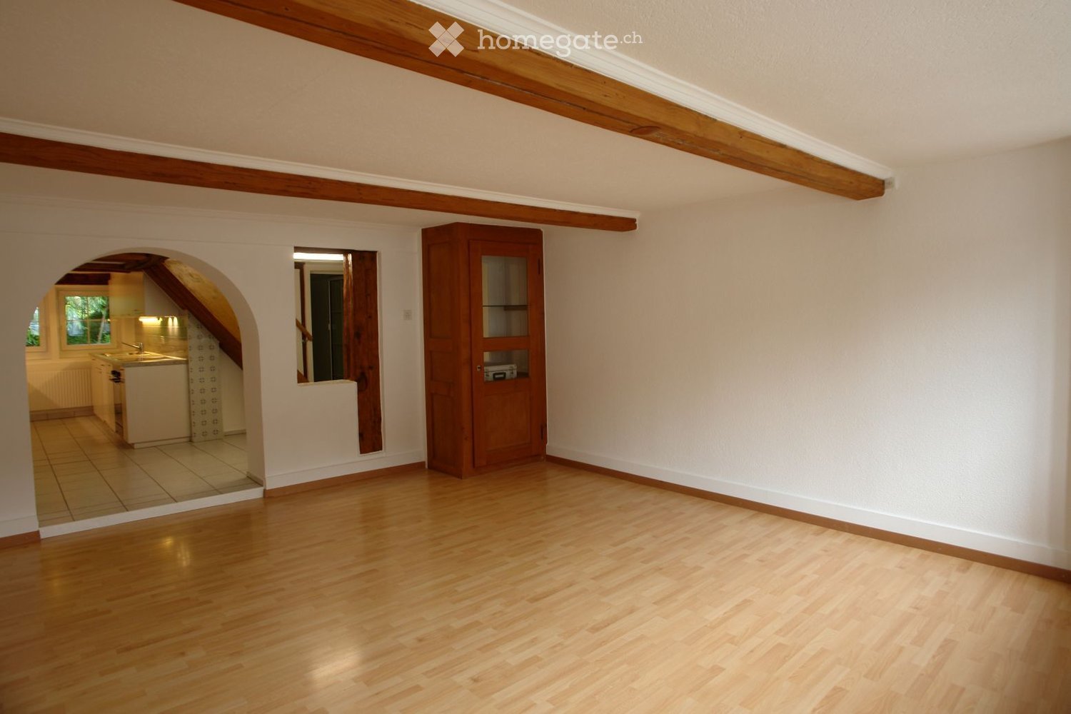 Empty room with wooden flooring, white walls, and exposed wooden beams on the ceiling. An arched doorway leads to a kitchen area with white cabinets and a sink.