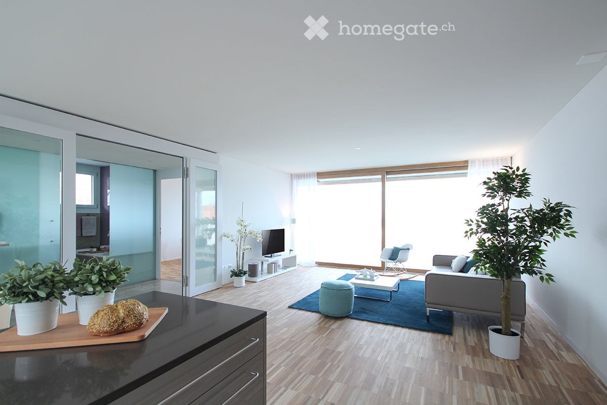 Bright living room with wooden flooring, large windows, grey sofa, coffee table, ottoman, armchair, television, and potted plants.