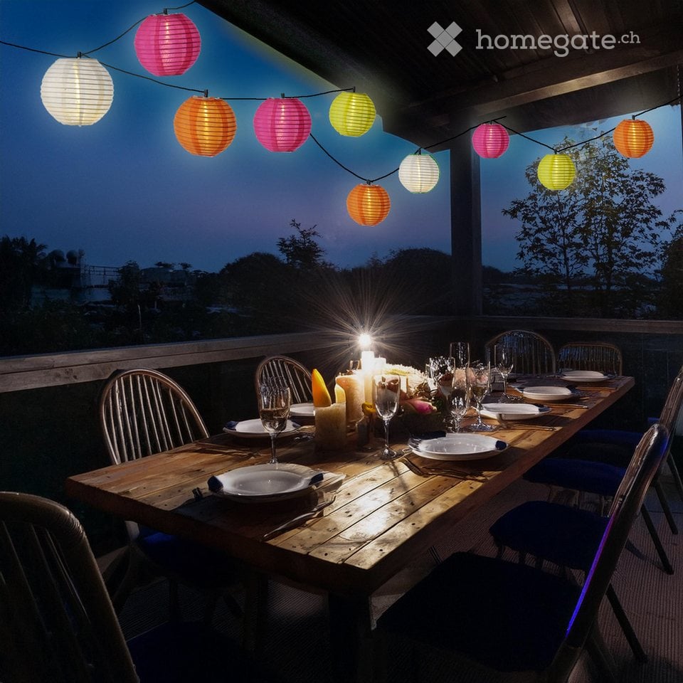 A wooden dining table set for a meal with plates, glasses, and candles, surrounded by colorful paper lanterns and blue chairs, set against a twilight backdrop with trees.