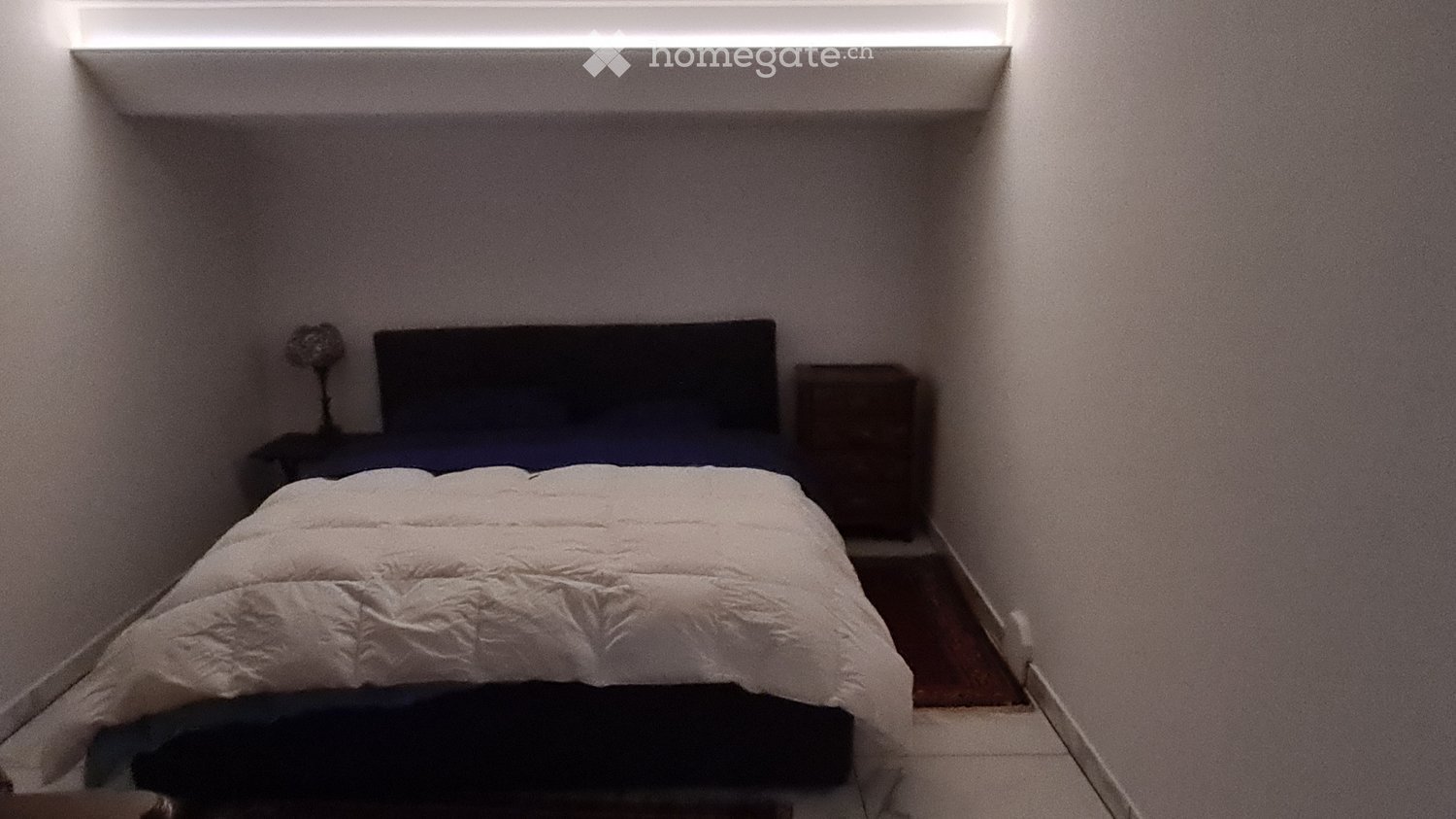 Small bedroom with a neatly made bed, a nightstand, and a lamp. The room has white walls and a built-in shelf above the bed.