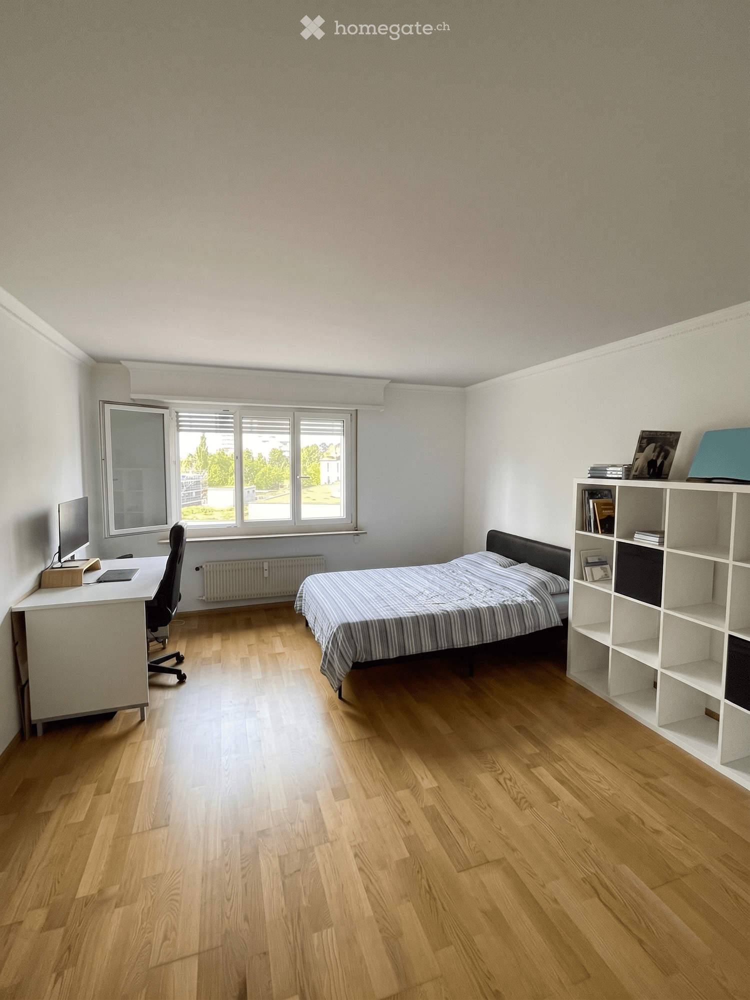 A furnished bedroom with a bed, desk, chair, shelf, and screen. The room features wooden flooring and large windows with blinds.