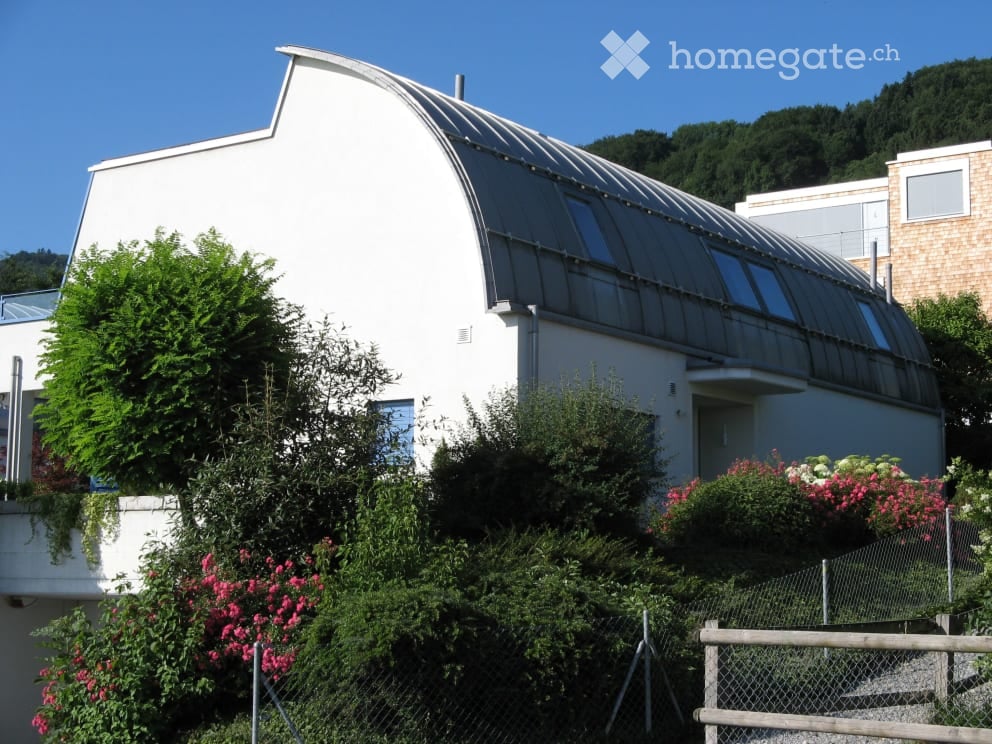 Modern white building with a curved roof and large glass panels, surrounded by lush greenery and flowering plants.