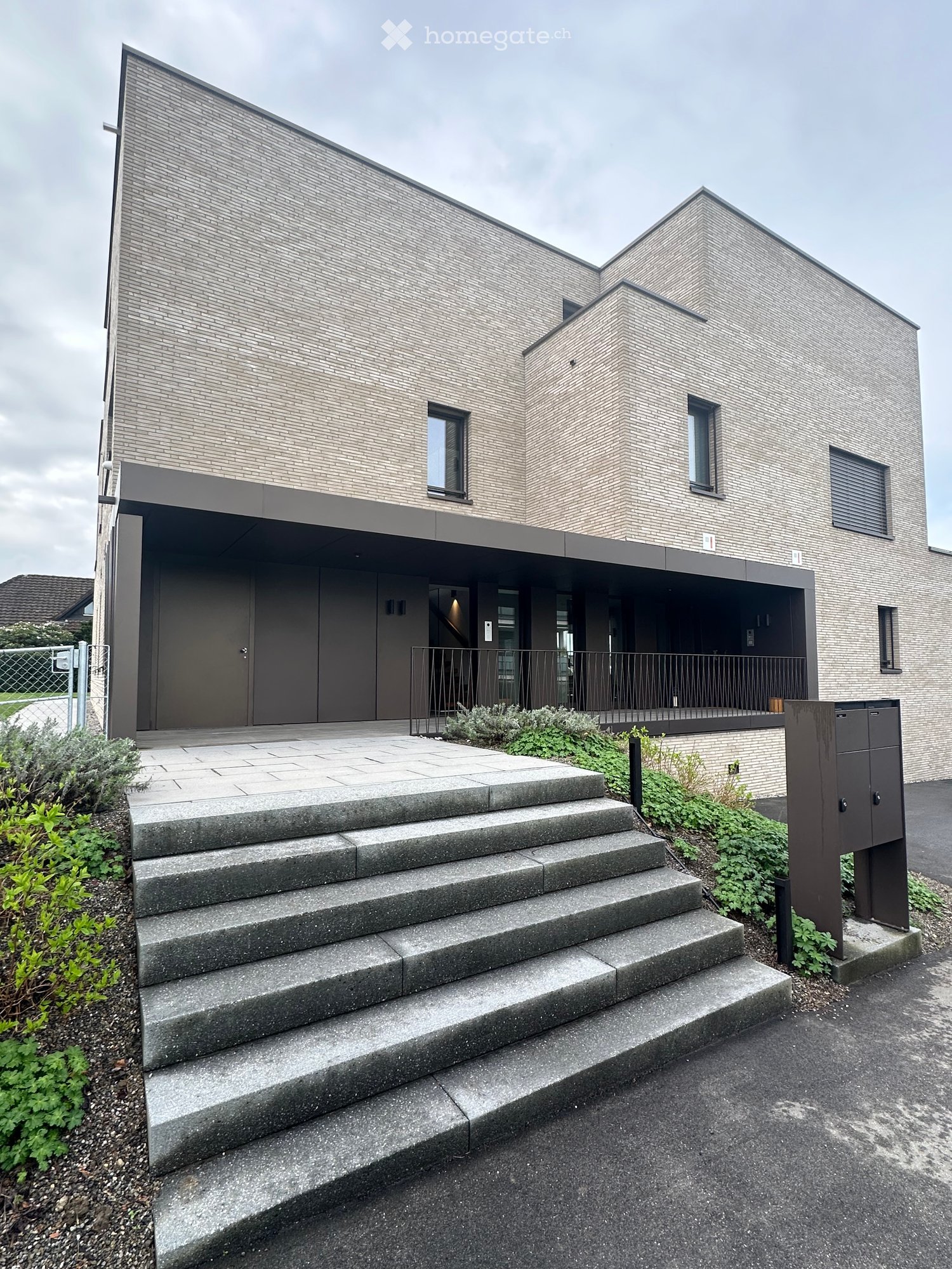 Modern two-story house with a brick facade, large windows, and a covered entrance area with steps leading up to the door.