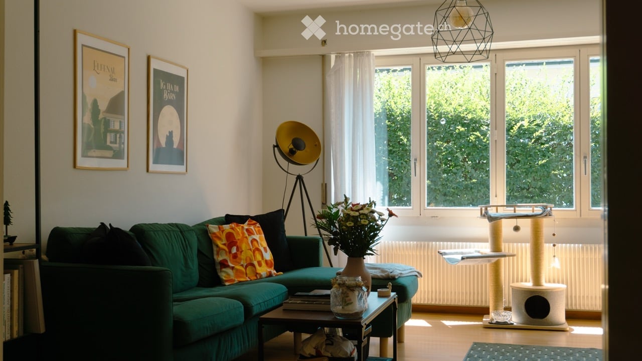 A modern living room with a green sofa, colorful throw pillow, coffee table with books and a vase of flowers, cat tree and cat bed, large windows with white curtains, and decorative wall art.