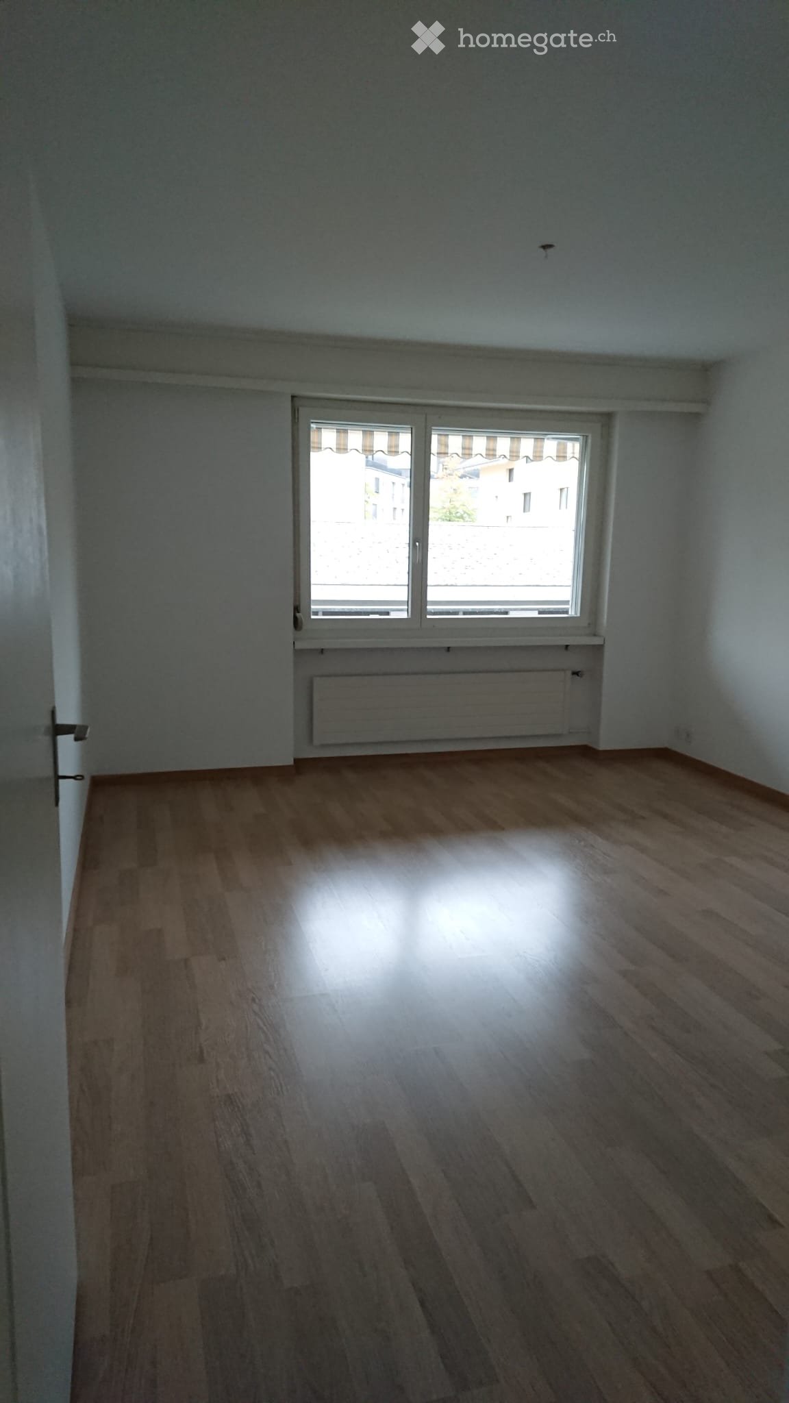 Empty room with wooden flooring, white walls, and a large window with blinds. A radiator is visible below the window.