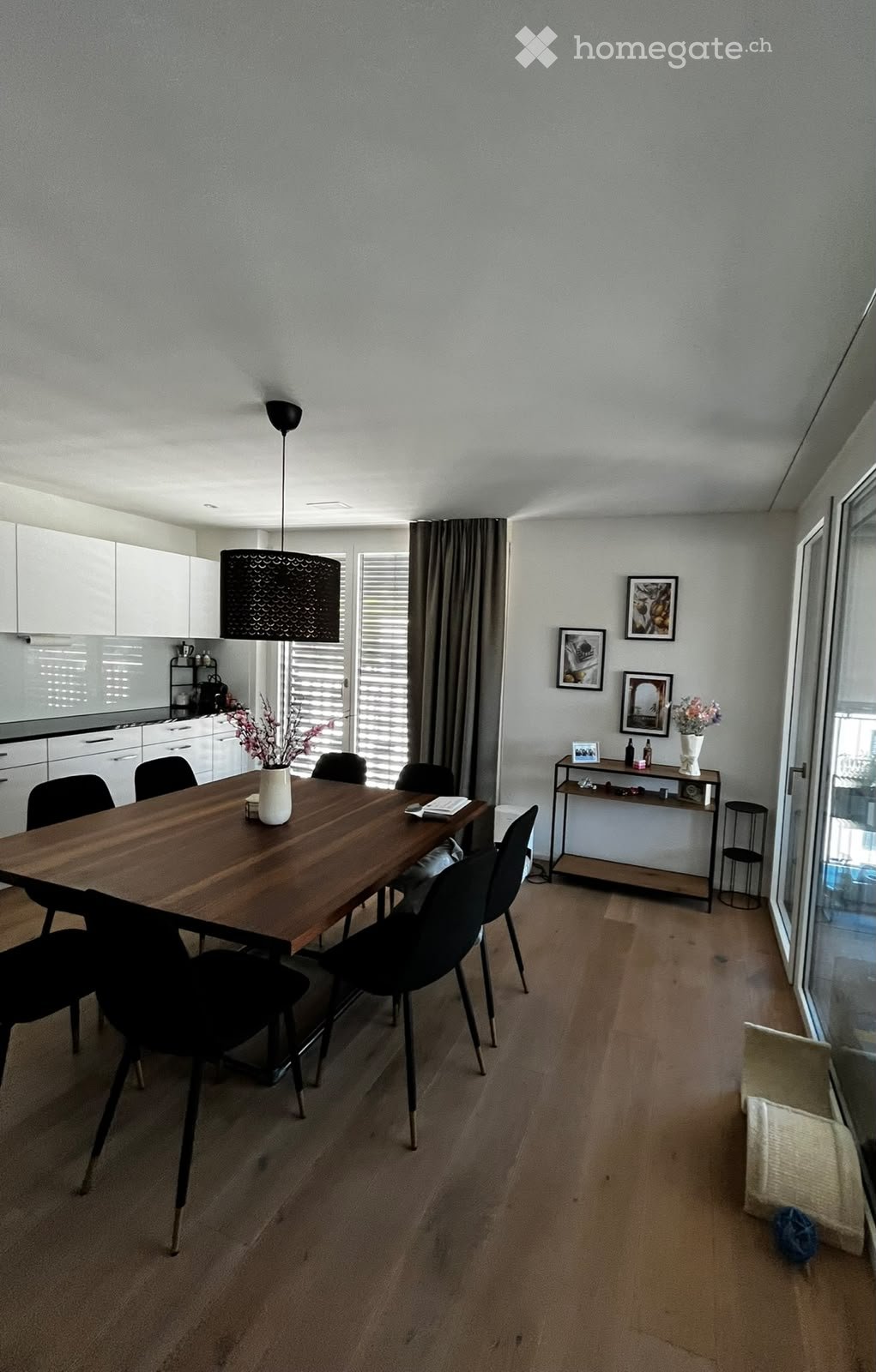 Modern dining area with a wooden dining table, black chairs, pendant light, white kitchen cabinets, and a small shelf with decorative items.