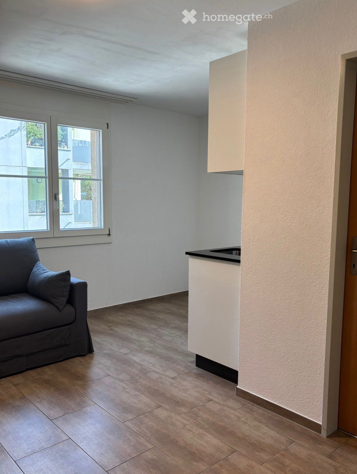 A modern, open-plan living and kitchen area with a dark grey sofa, a white kitchen counter, and large windows offering a view of the outside.