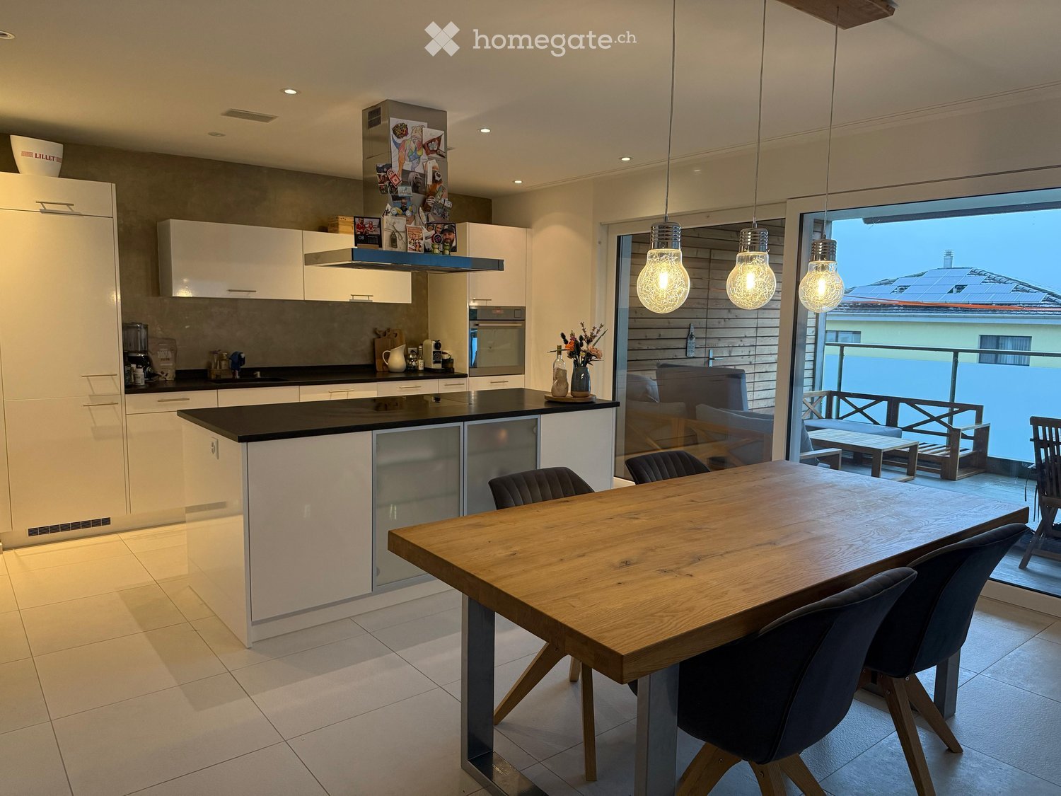 Modern kitchen with white cabinets, black countertop, island, dining table, and pendant lights. Offers a view of the outdoor area through large windows.