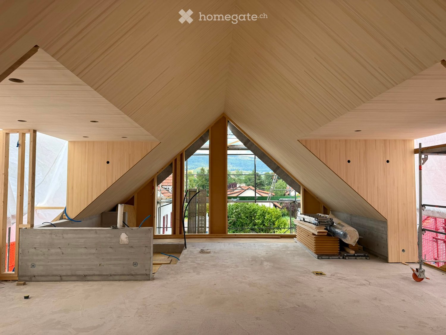 An attic space with wooden walls and ceiling, large windows offering a view, and construction materials stacked in the corner.