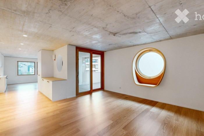 Empty room with wooden flooring, white walls, and large windows, including a round porthole window and a sliding glass door leading to another space.