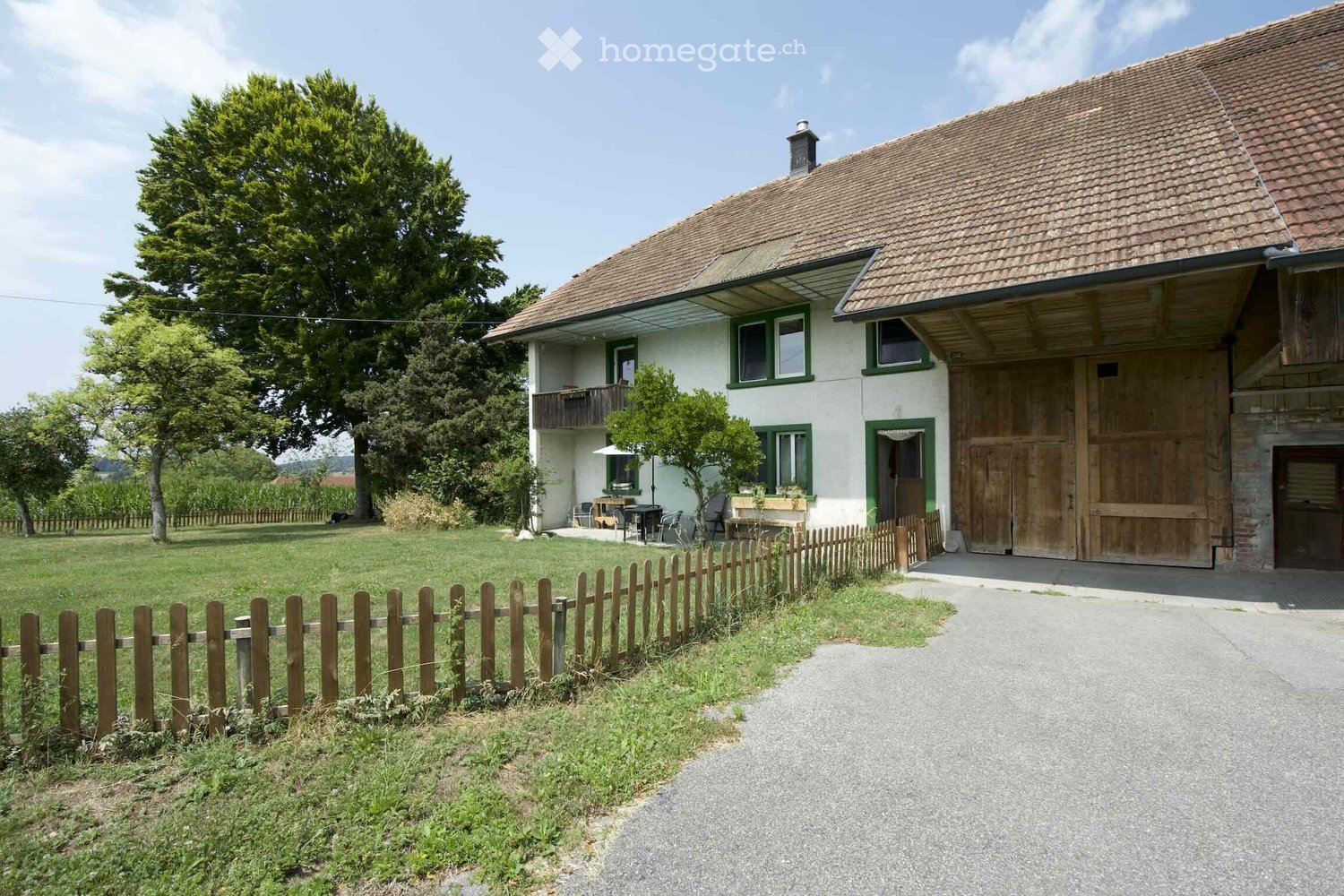 Two-story house with a tiled roof, white walls, green-framed windows, and a wooden garage. The house is surrounded by a wooden fence, a lawn, and trees.