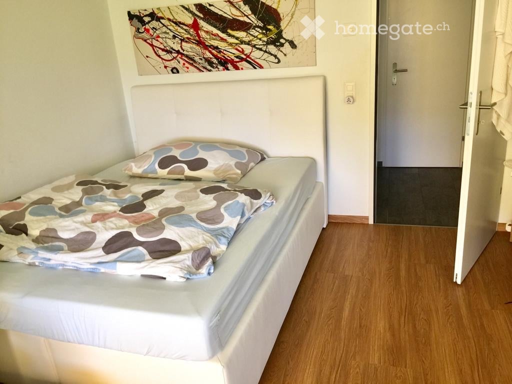 A bed with white mattress, wooden floor, white headboard, bedspread with abstract patterns, pillow, entrance door