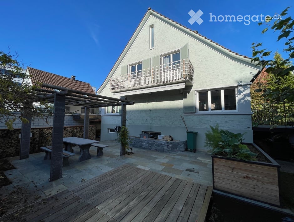 Outdoor patio area with wooden deck, stone seating area, and a two-story house with a balcony in the background.