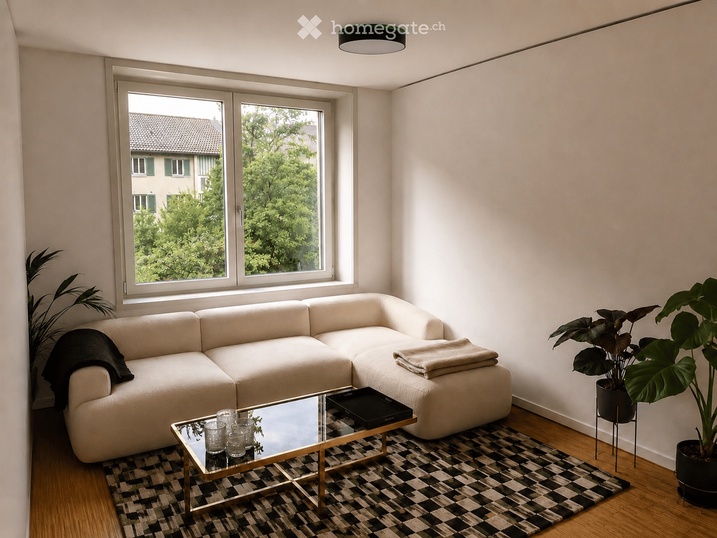 Modern living room with a light-colored sectional sofa, glass coffee table, and potted plants. The room features large windows with a view of greenery outside, a patterned rug, and a sleek ceiling light fixture.