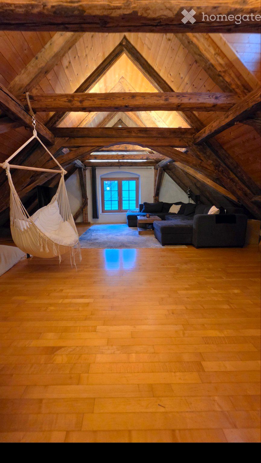 Attic living space with wooden beams, exposed wooden ceiling, hardwood floor, large window, hammock, sofa, coffee table, and ottoman.
