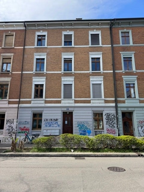 Four-story brick building with multiple windows, graffiti on the lower facade, and a small garden area in front.