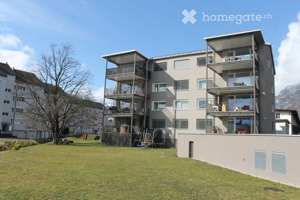 Modern multi-story apartment building with multiple balconies, large windows, and a view of mountains in the background. The building is surrounded by a grassy area with playground equipment.