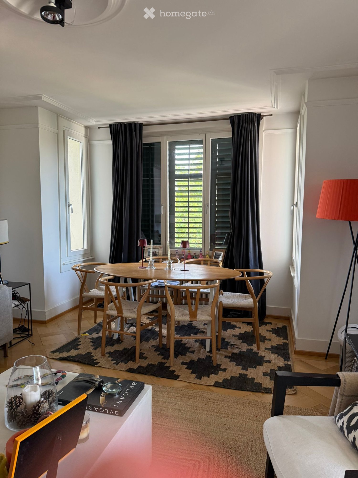 A modern, well-lit living and dining area with a round wooden dining table, six chairs, a sofa, a floor lamp, and decorative items on a side table.