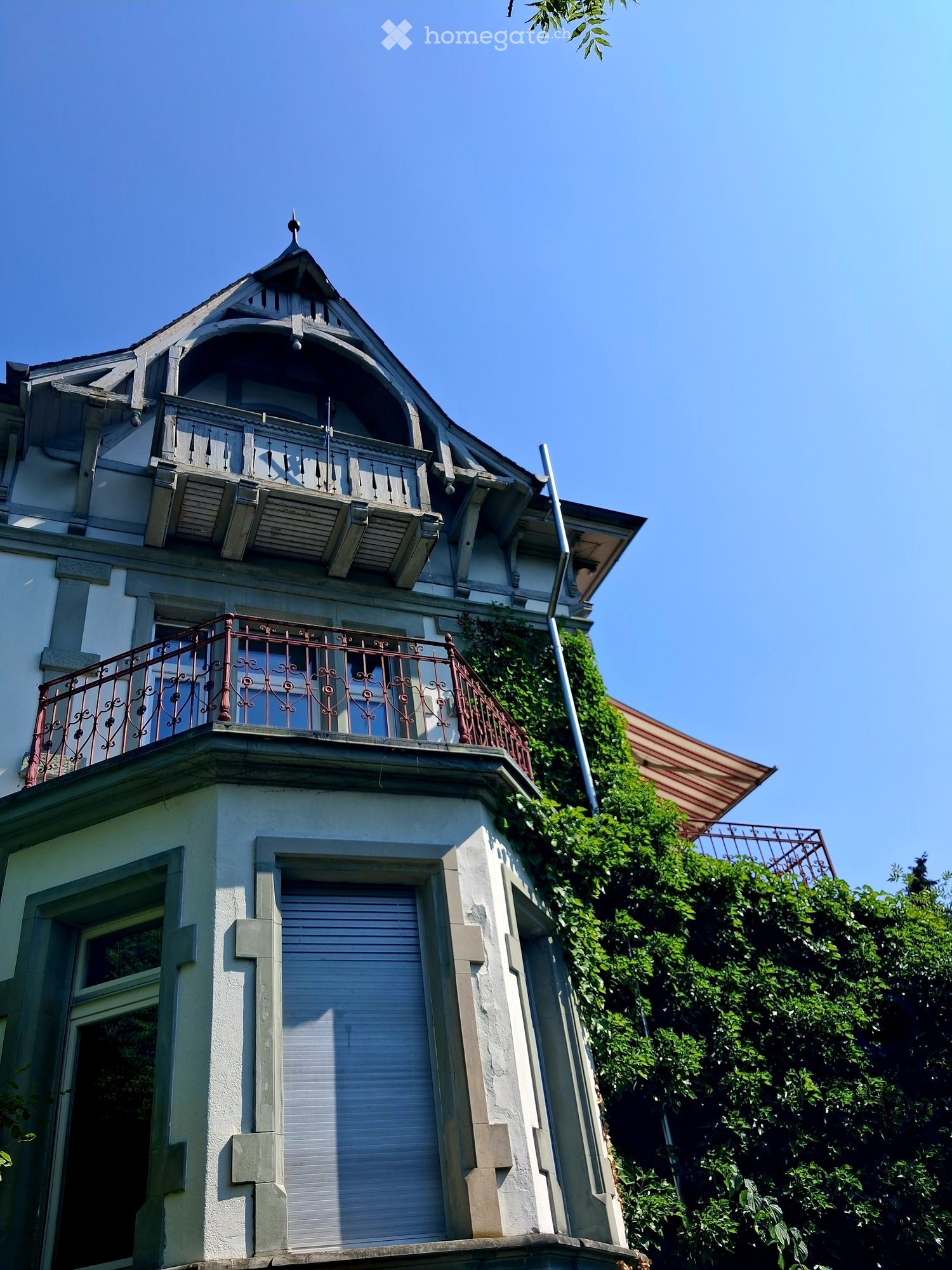 A multi-story residential building with ornate architectural details, including a decorative balcony with wrought-iron railings, a small turret with a pointed roof, and a covered entrance with a shuttered door.