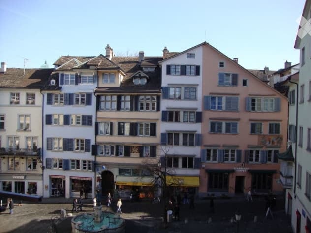 A row of multi-story buildings with classic European architecture, featuring multiple windows, shutters, and a central square with a fountain and people walking.