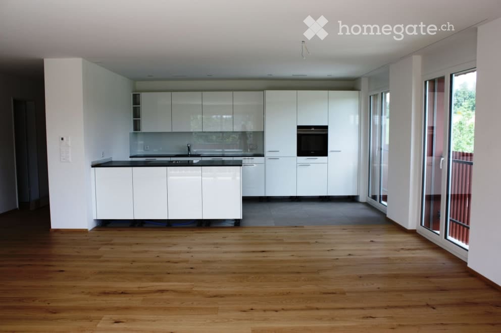 Open-plan kitchen and living area with white cabinetry, dark countertops, and wooden flooring. Large windows and sliding glass doors lead to an outdoor space.
