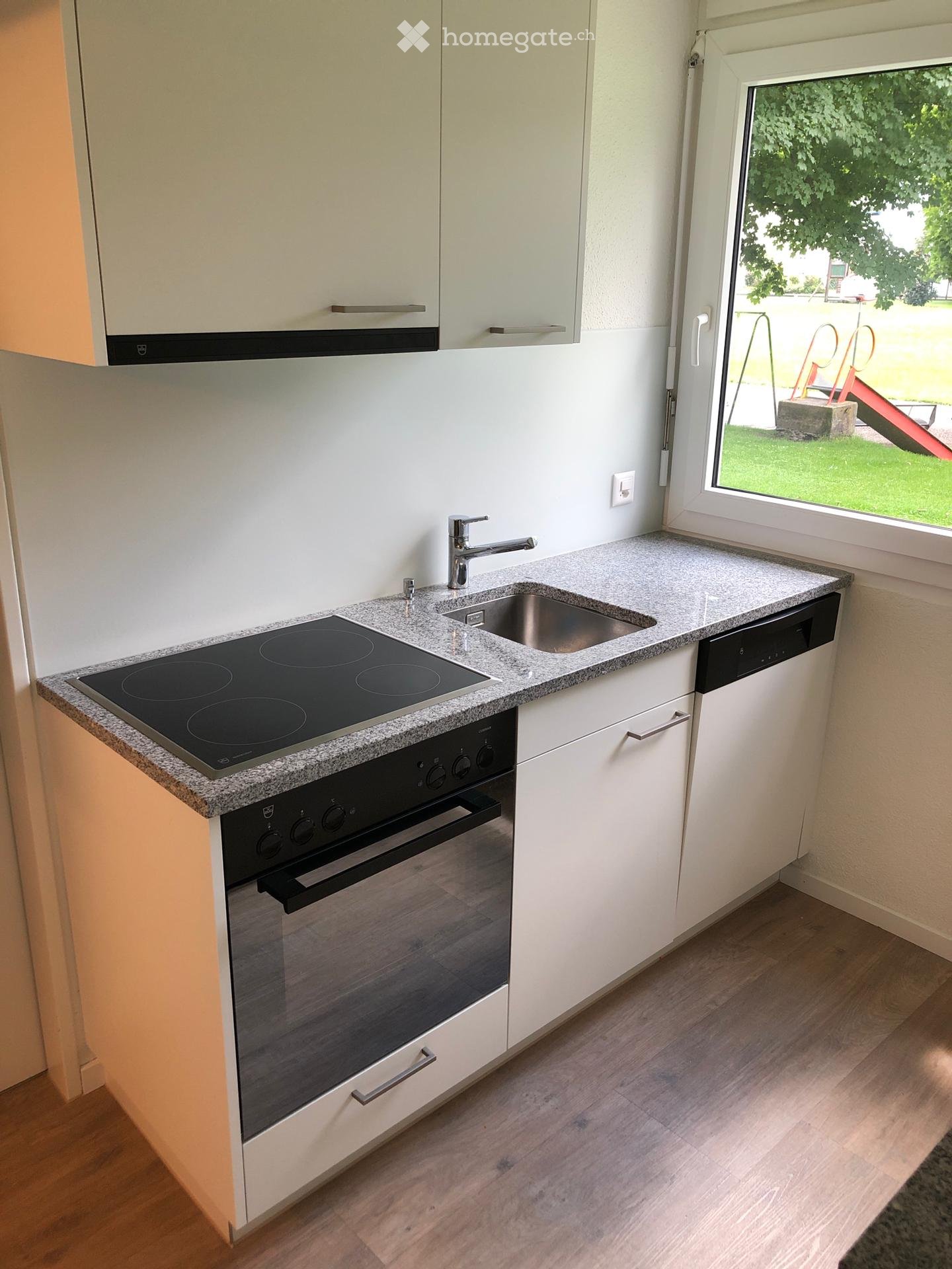 Modern kitchen with white cabinets, granite countertop, induction stove, oven, sink, and dishwasher. Large window offers view of outdoor area.