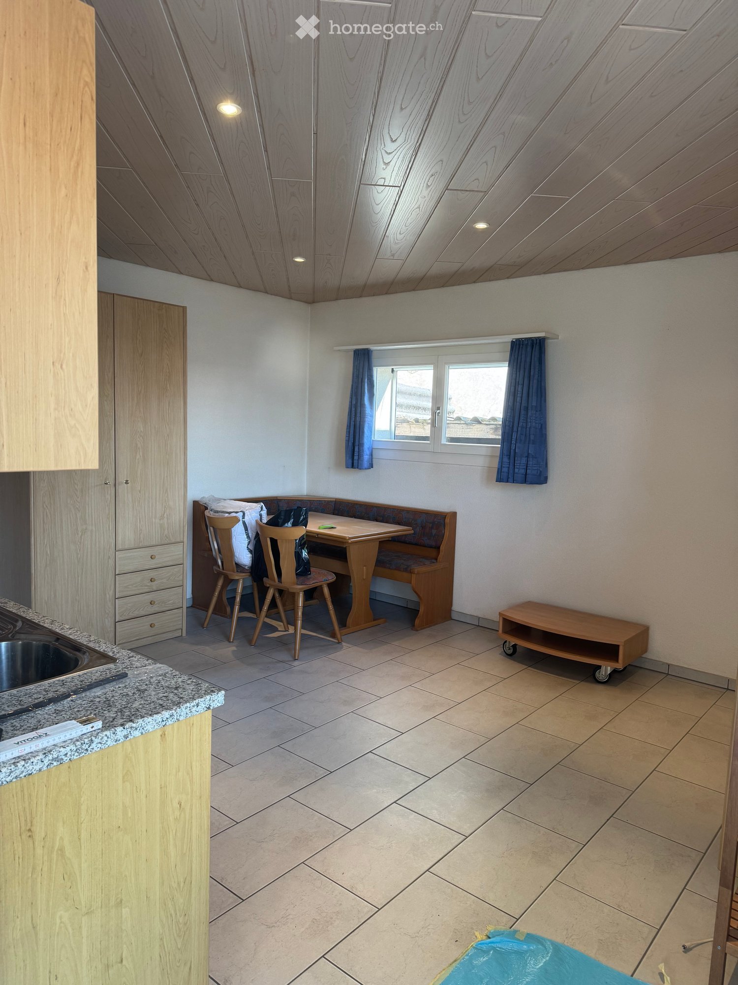 kitchen with dining area, wooden furniture, white walls, tiled floor, window with blue curtains