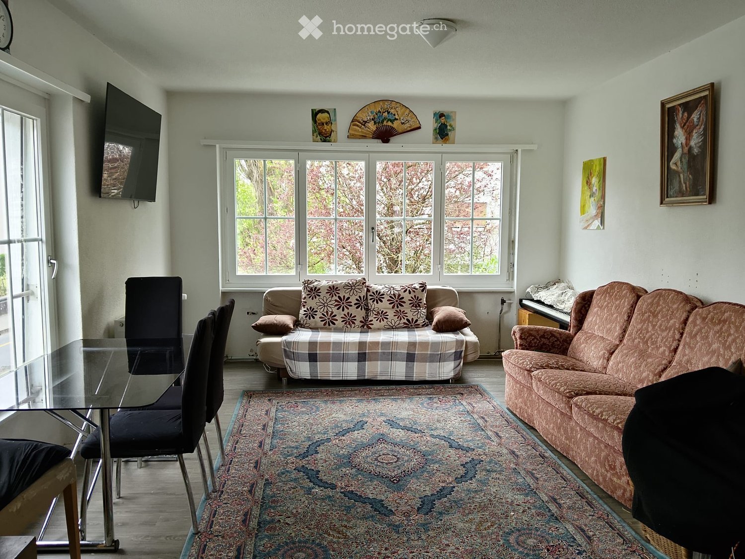 Bright living room with large windows, two sofas, a glass dining table with chairs, a piano, and a television mounted on the wall.