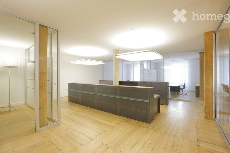 Modern office space with a reception desk, chairs, and a table, featuring large glass walls and a polished wooden floor.