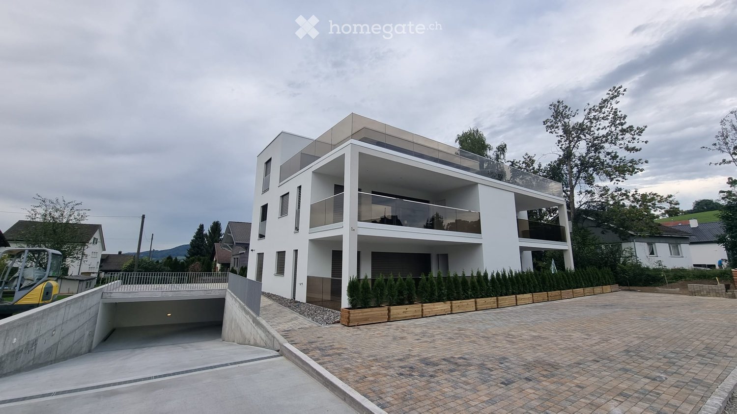 Modern two-story house with white facade, large windows, and balconies. Features a covered parking area and landscaped entrance.