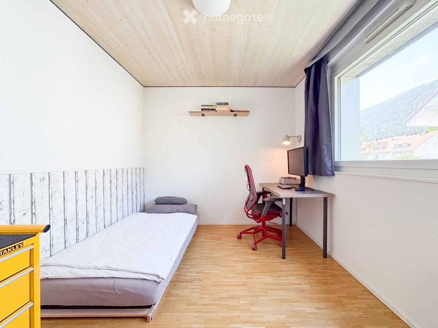 A small, modern bedroom with a single bed, a desk with a chair, a wall-mounted shelf with books, and a window with a view of the mountains.