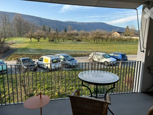 Balcony with a view of a green field, parking area, and distant mountains. Features a round table and a wicker chair.