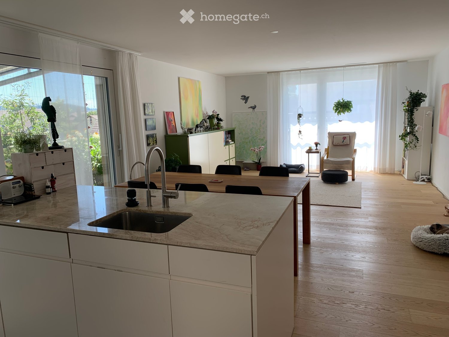 Open-plan kitchen and living area with a central island, sink, and seating area. The space features light wooden flooring, large windows with sheer curtains, and modern furniture including a dining table, chairs, and a small living area with a stool and d