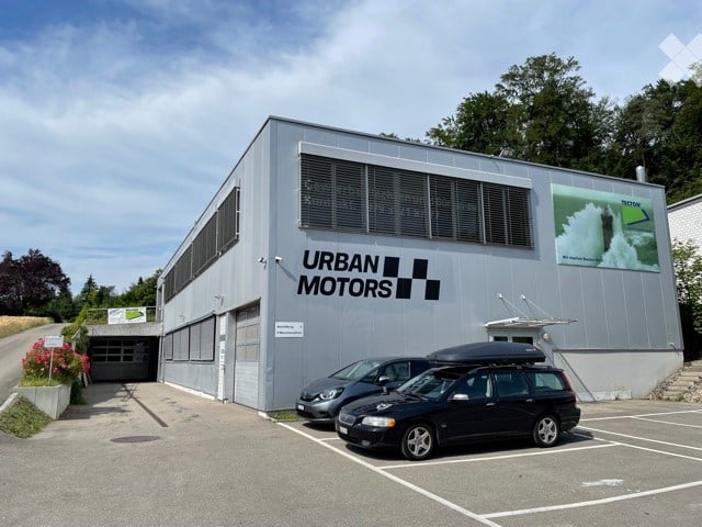 Modern single-story building labeled "URBAN MOTORS" with large garage doors, several parked cars, and a small landscaped area with flowers.